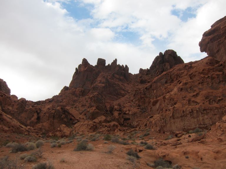 looking up at red rock cliffs against a blue but cloudy sky with green shrubs in the foreground