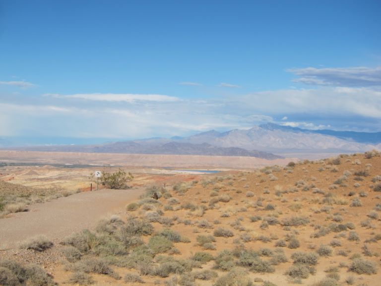 a gravel road with a faint no parking sign in the distance to the left surrounding the road is a broan desert landscape with lots of green bushes and mountains in the background against a blue sky with clouds